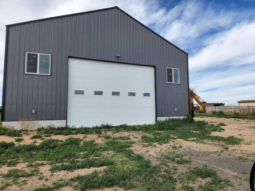 Metal barn with large garage door, surrounded by overgrown grass and machinery in background.